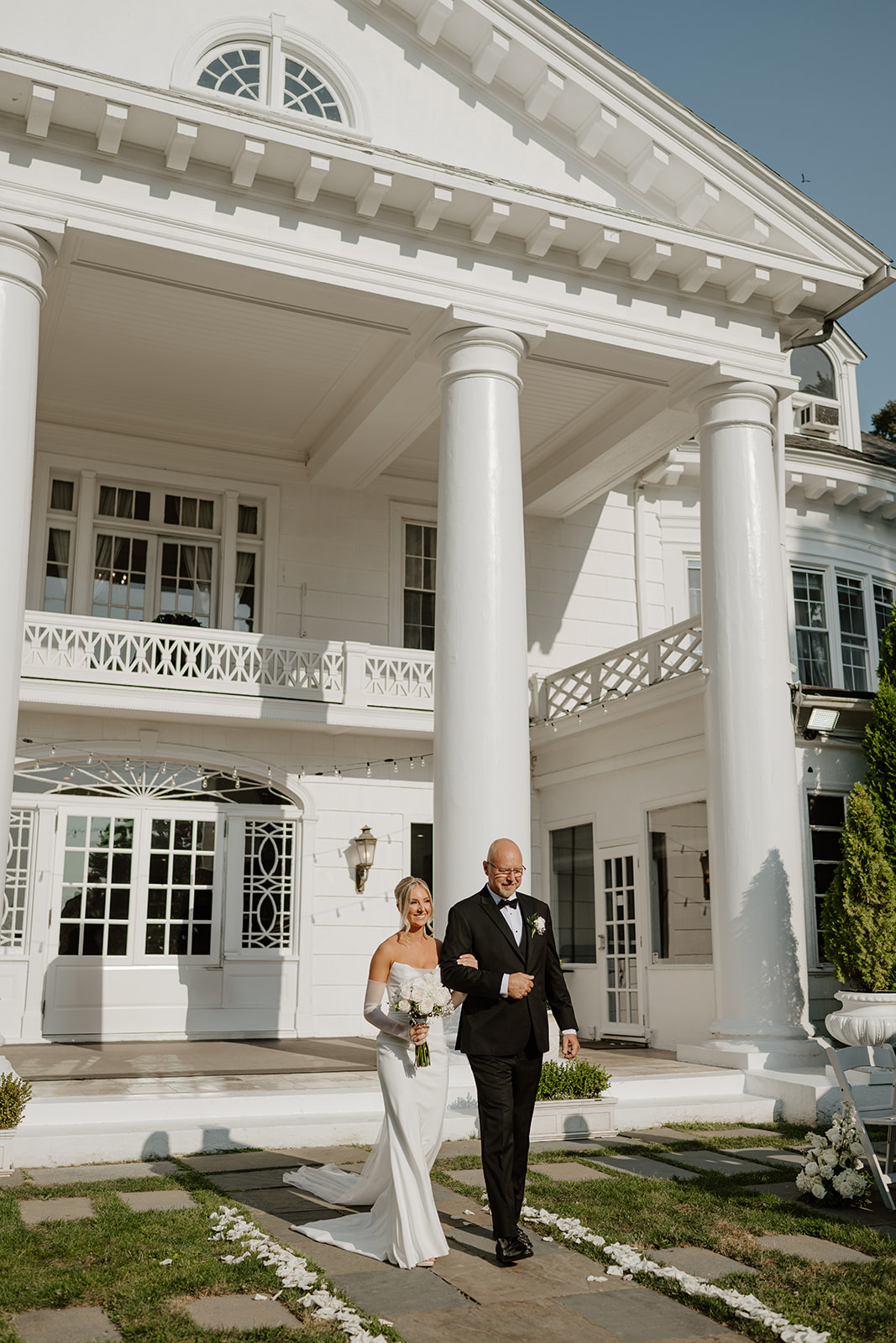 Smiling bride walks arm-in-arm with her father down the aisle outside a grand columned mansion.